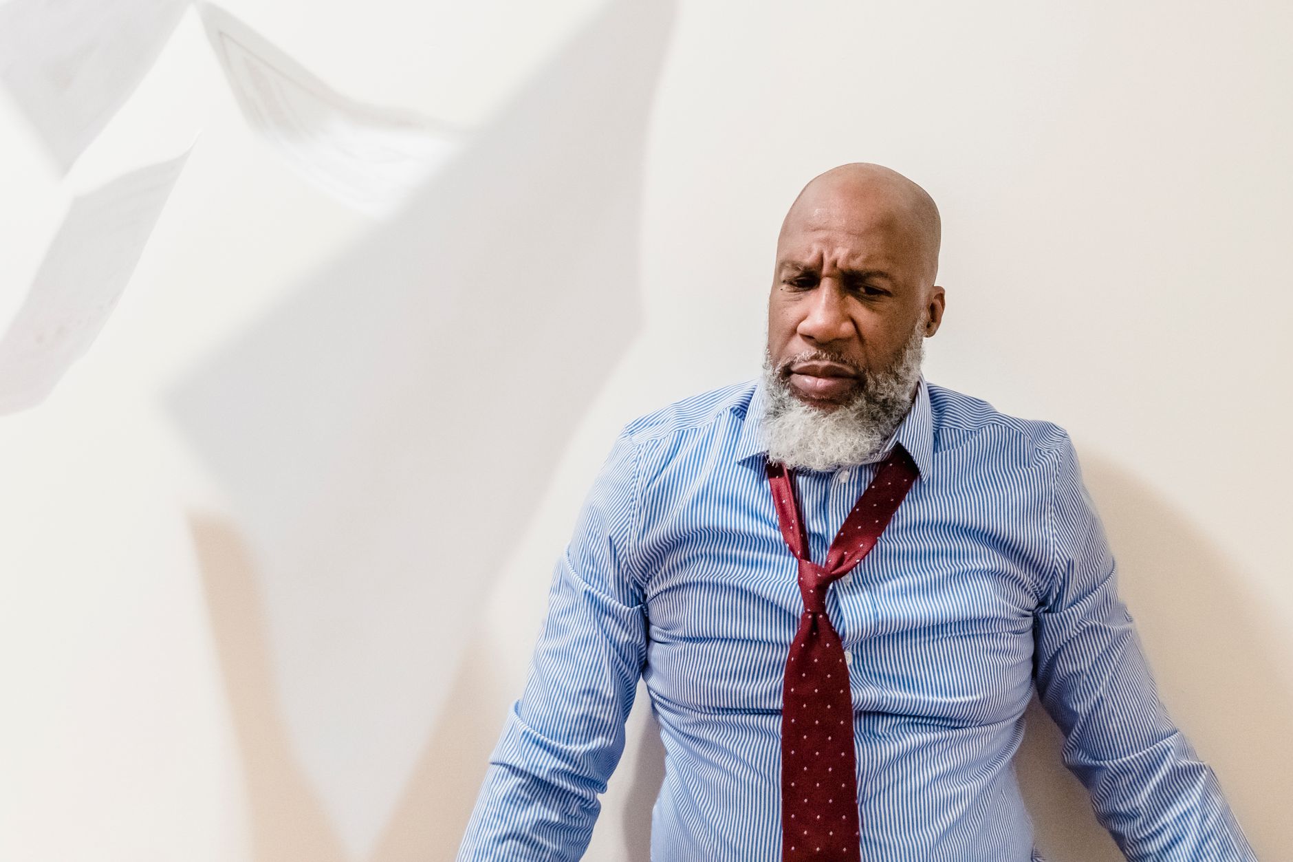 A man in a shirt and tie looks frustrated as papers fall around him indoors.