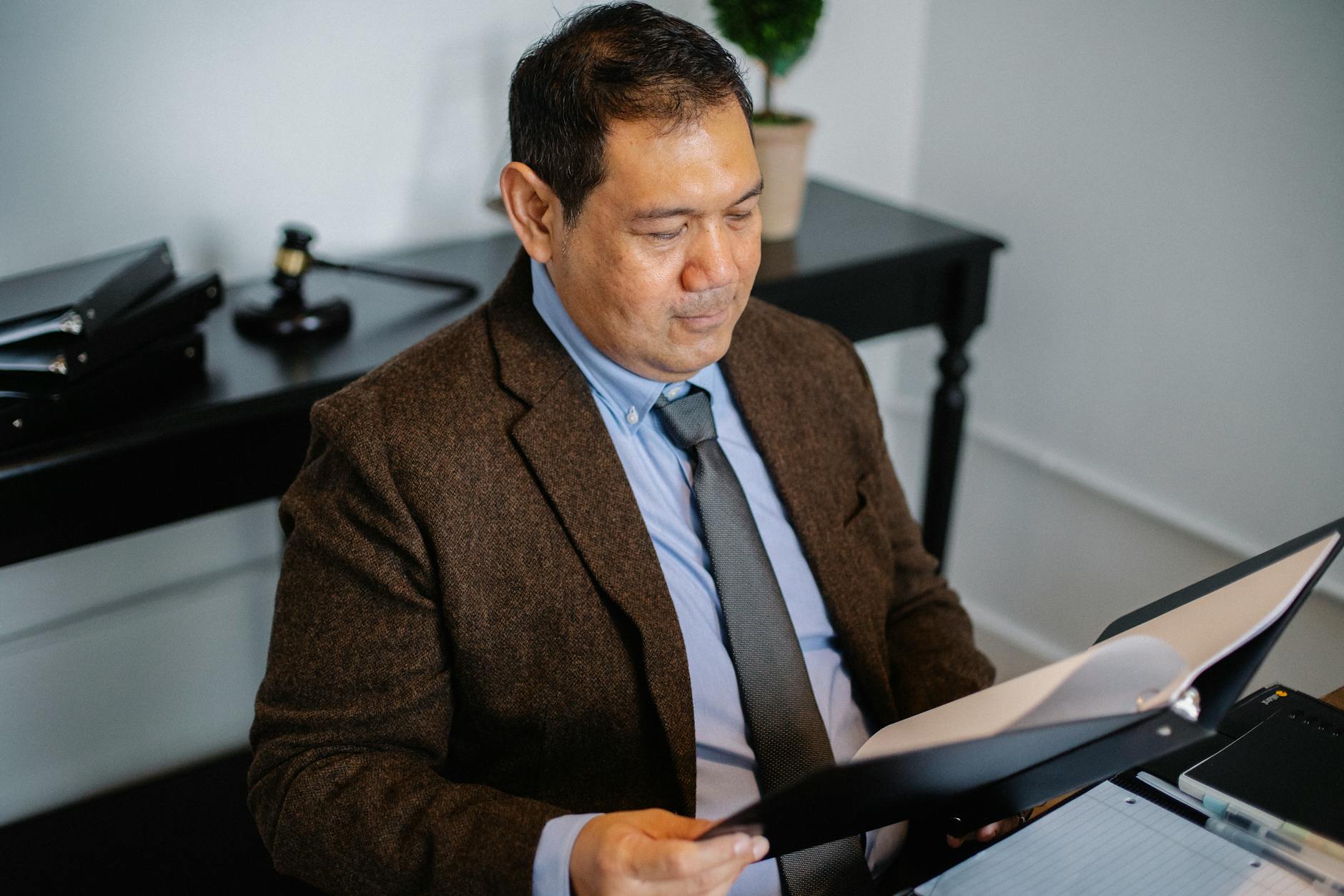 From above of serious ethnic lawyer in elegant suit checking report in folder on blurred background of office.